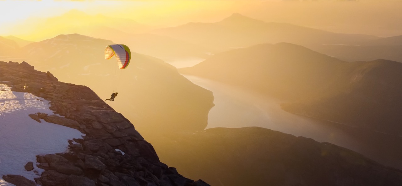 Ein Gleitschirmflieger startet im Abendlicht von einer Felskante, darunter ein Fjord zwischen Bergen