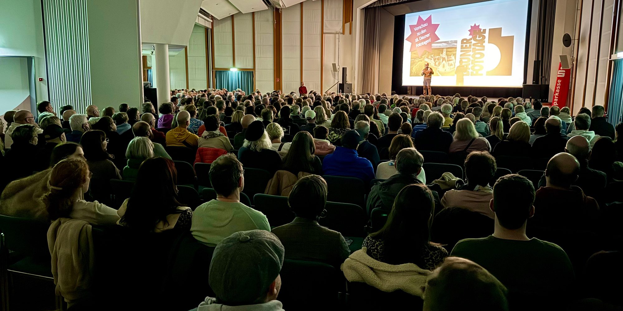 Voll besetzter Saal im Forum Daun, das Publikum von hinten, vorn die Leinwand und der Moderator auf der Bühne