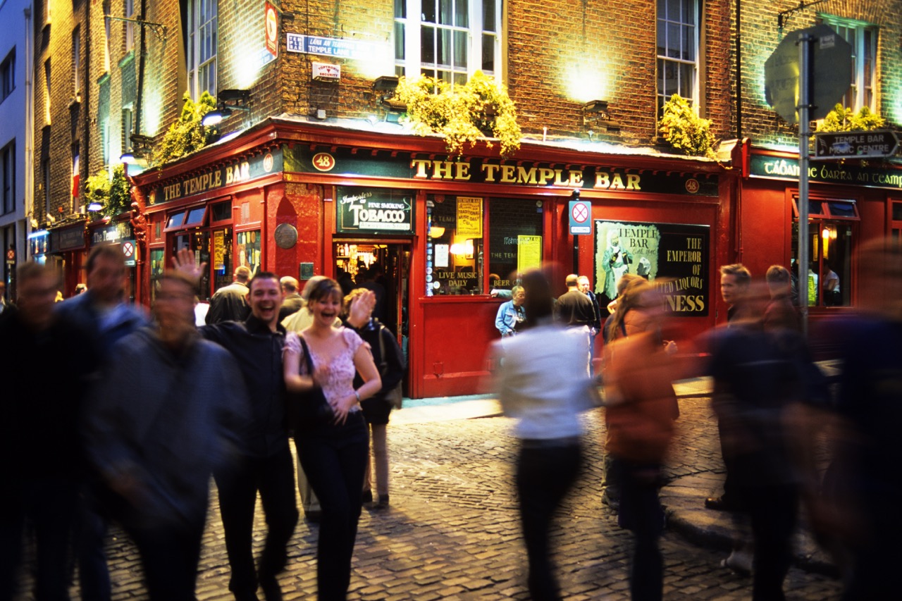 Abendliche Straßenszene vor der roten Fassade des Pubs The Temple Bar in Dublin, Passanten teils in Bewegungsunschärfe