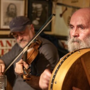 Zwei Musiker im Pub, vorn ein Mann mit Bart und Rahmentrommel, dahinter ein Geiger mit Schirmmütze