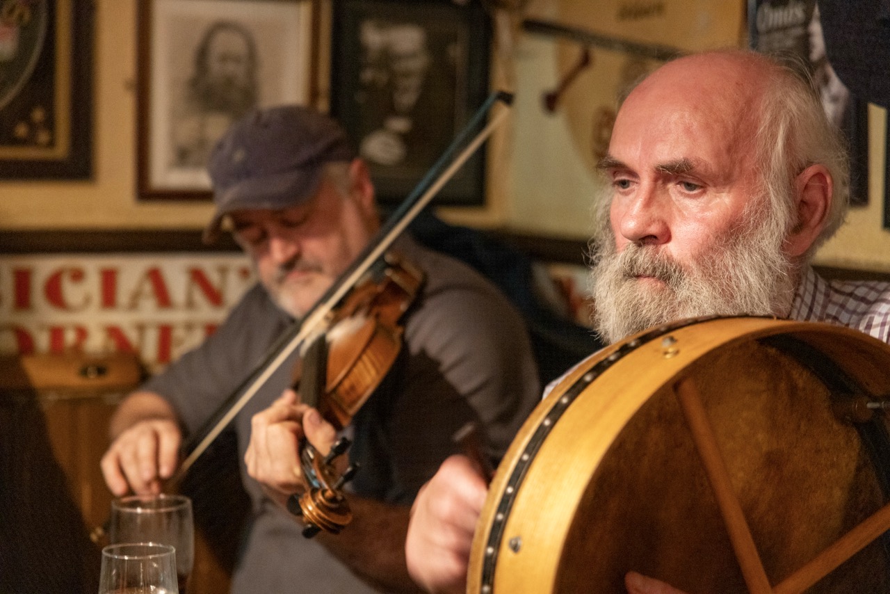Zwei Musiker im Pub, vorn ein Mann mit Bart und Rahmentrommel, dahinter ein Geiger mit Schirmmütze