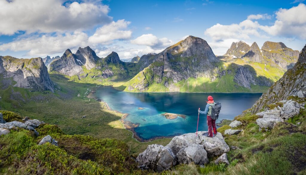 Wanderin mit rotem Hut auf einem Felsen über einem türkisfarbenen Fjord, ringsum steile Berge